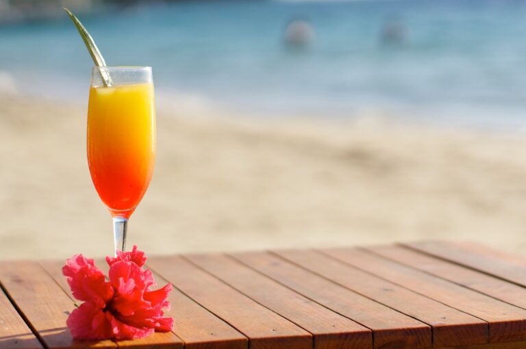 A colorful tropical cocktail with a pineapple garnish is placed on a wooden table with pink flowers in front, overlooking the ocean in Roatan, Honduras.