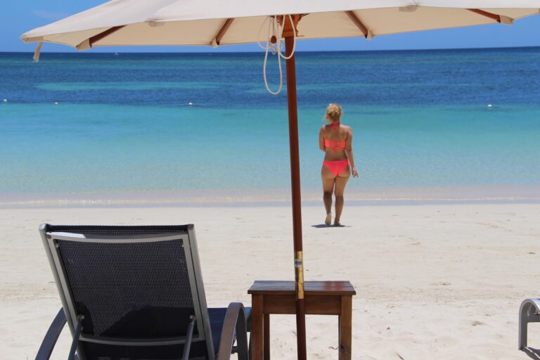 A woman in a coral bikini walks along the sandy beach in Roatan, Honduras, under a large white beach umbrella with the turquoise ocean and blue sky in the background.