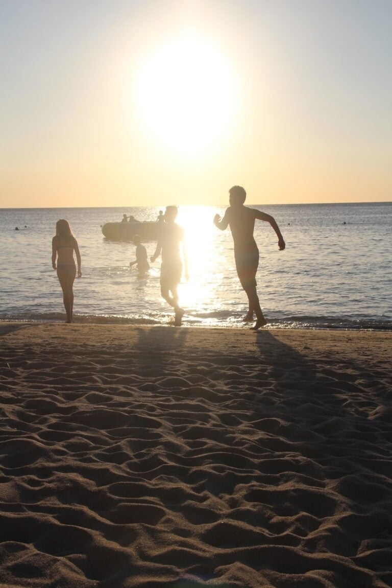 People enjoy a sunset on the beach in Roatan, Honduras, with silhouettes of individuals walking and swimming in the calm ocean water against a bright, glowing sun.