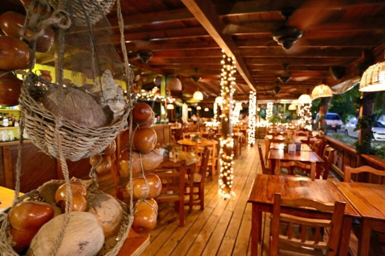 A cozy wooden restaurant in Roatan, Honduras, decorated with string lights and wicker lampshades, features a basket of onions hanging near the ceiling.