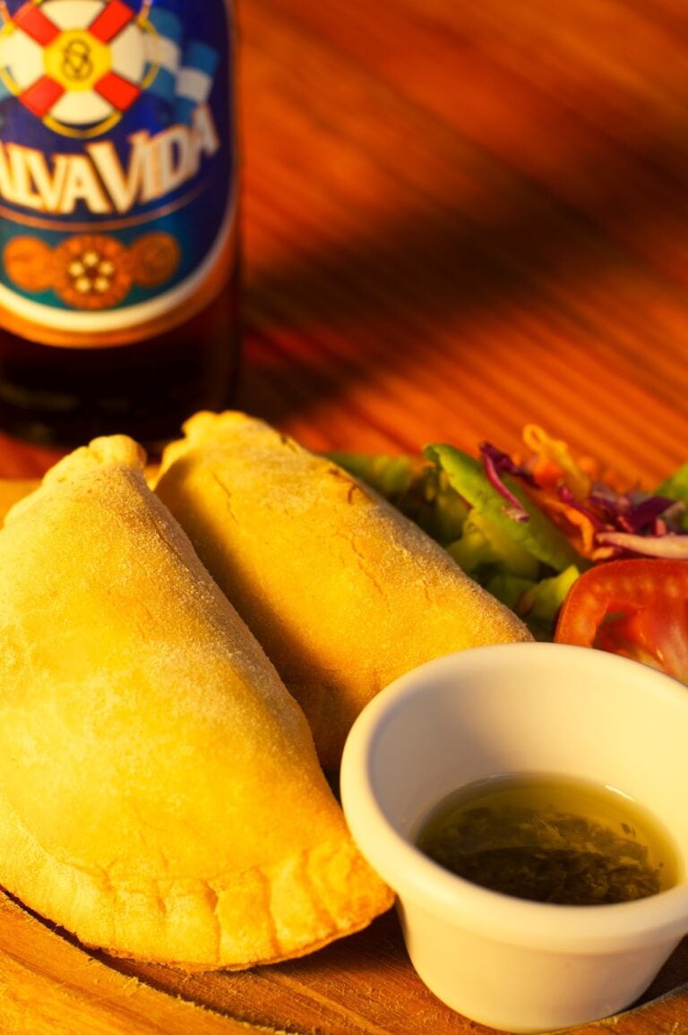 A close-up of two crispy, golden fried Colombian arepas with a small white cup of green herb sauce and colorful grilled vegetables on a wooden platter in Roatan, Honduras.