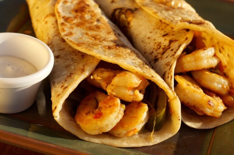 Close-up of three grilled tacos filled with shrimp and vegetables, served with a side of creamy white sauce on a black plate in Roatan, Honduras.