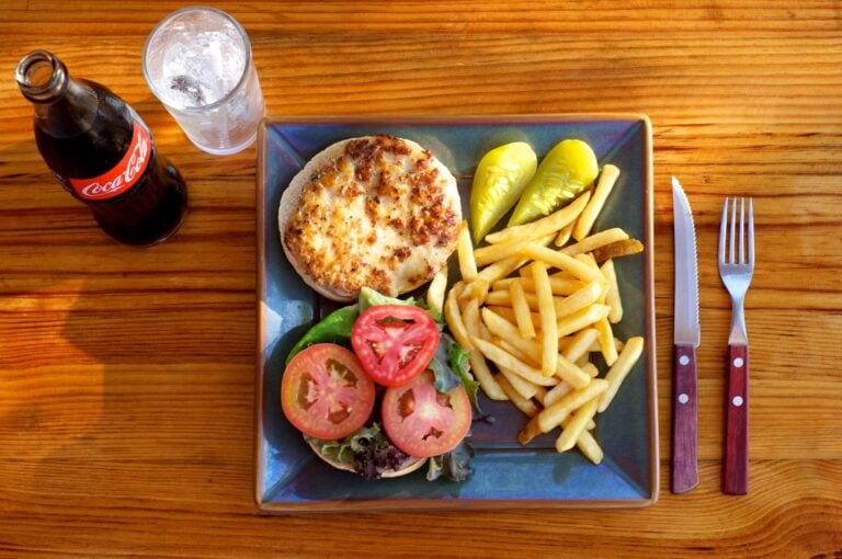 A plate with a cheeseburger, French fries, sliced tomatoes, lettuce, and pickles is shown on a wooden table along with a glass of water, a Coca-Cola bottle, and a fork and knife, representing casual dining in Roatan, Honduras.