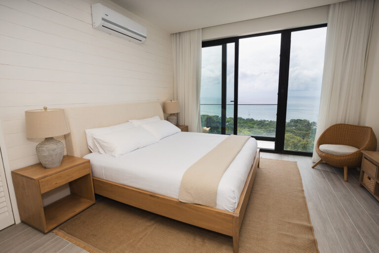 A bedroom in a Roatan, Honduras hotel features a large bed with white linens, two matching bedside tables with lamps, and a wicker chair near floor-to-ceiling windows overlooking the ocean and lush greenery.