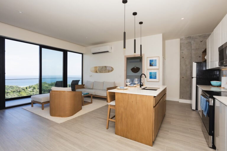 A modern, open-concept living and kitchen area in a residential property on Roatan, Honduras, featuring large glass doors with ocean views, a beige sofa, wooden dining table with wine glasses, contemporary pendant lighting, and a mix of concrete and wooden interior design elements.