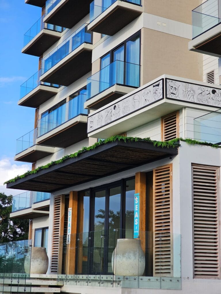 A modern multi-story building in Roatan, Honduras, features glass balconies, wooden accents, greenery on the terrace, and decorative artwork on the exterior wall.