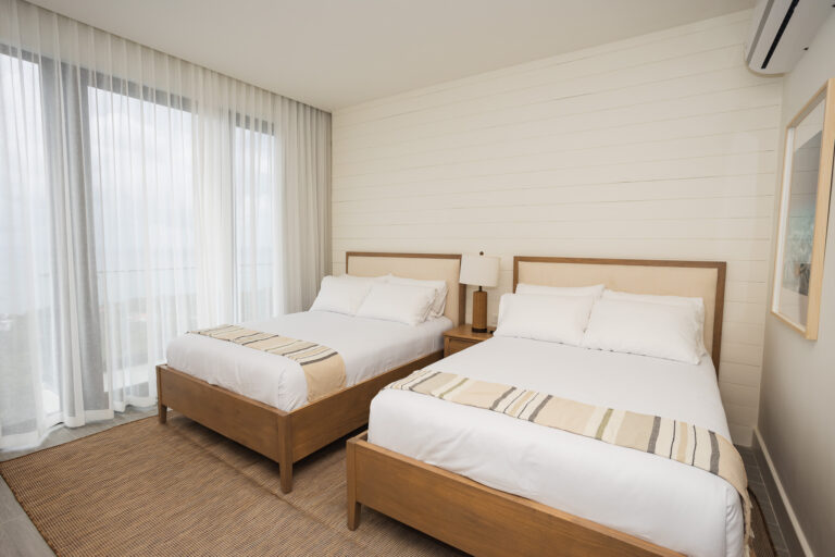 A modern hotel room in Roatan, Honduras, features two neatly made beds with wooden frames, a bedside table with a lamp, large windows with sheer white curtains, and a wall-mounted air conditioning unit.