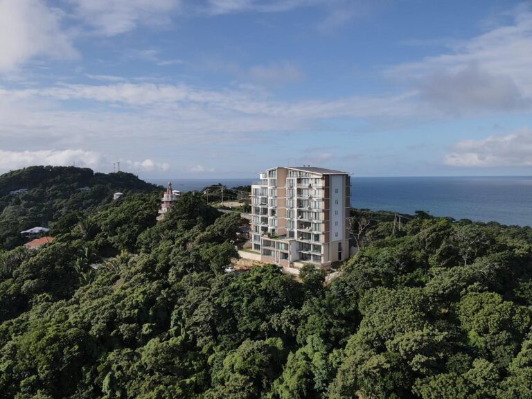 A tall modern apartment building overlooks a lush green hillside with trees and smaller houses, with the blue Caribbean Sea and a partly cloudy sky in the background, in Roatan, Honduras.
