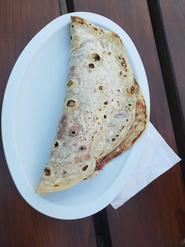 A large partially folded wrap sandwich with what appears to be ham and cheese filling served on a white oval plate, placed on a dark wooden table in Roatan, Honduras.