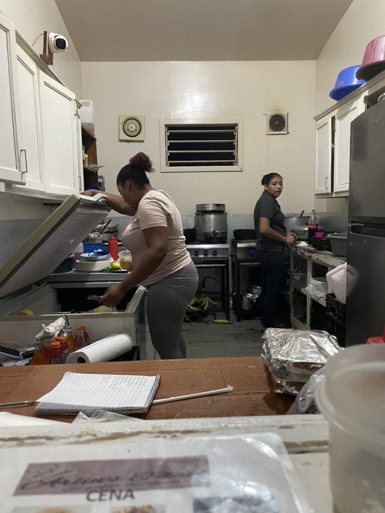 Two women are working in a small, busy kitchen in Roatan, Honduras, with white cabinets, a window with horizontal louvers, and various kitchen supplies, including condiments and food items on the counter.