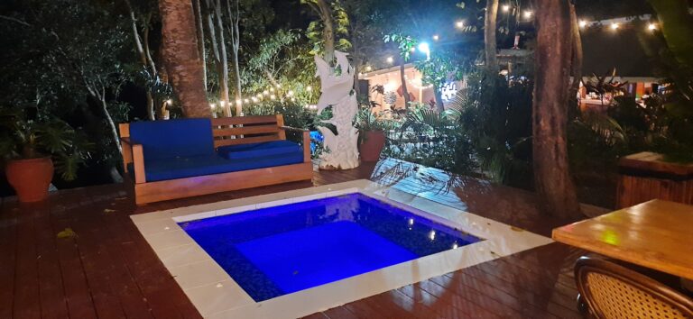 A cozy outdoor seating area with a small illuminated pool surrounded by lush tropical plants and string lights, located in Roatan, Honduras.