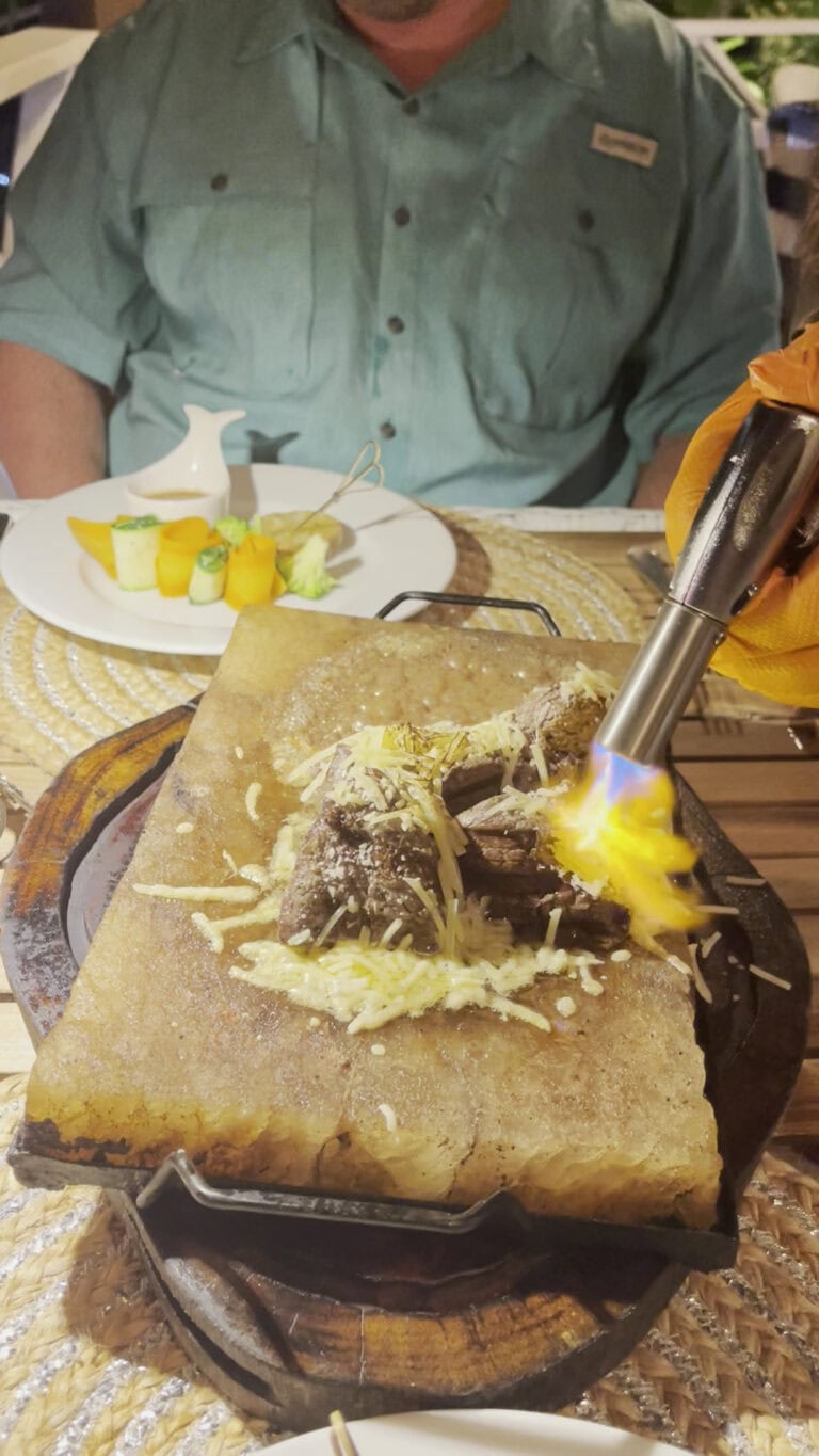 A person is preparing and serving traditional roasted meat on a wooden table in a restaurant setting in Roatan, Honduras, with a small plate of food and decorative elements visible in the background.