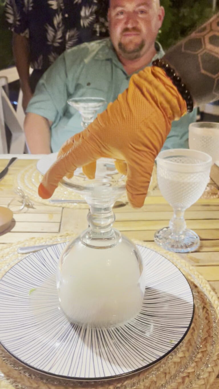 A person wearing an orange glove is holding a glass upside down on a table during a meal in Roatan, Honduras, with a man smiling in the background at an outdoor dining setting.