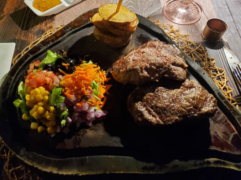 A sizzling platter of grilled meats accompanied by fresh salad, plantains, and sauces is displayed on a wooden table in a restaurant setting on Roatan, Honduras.