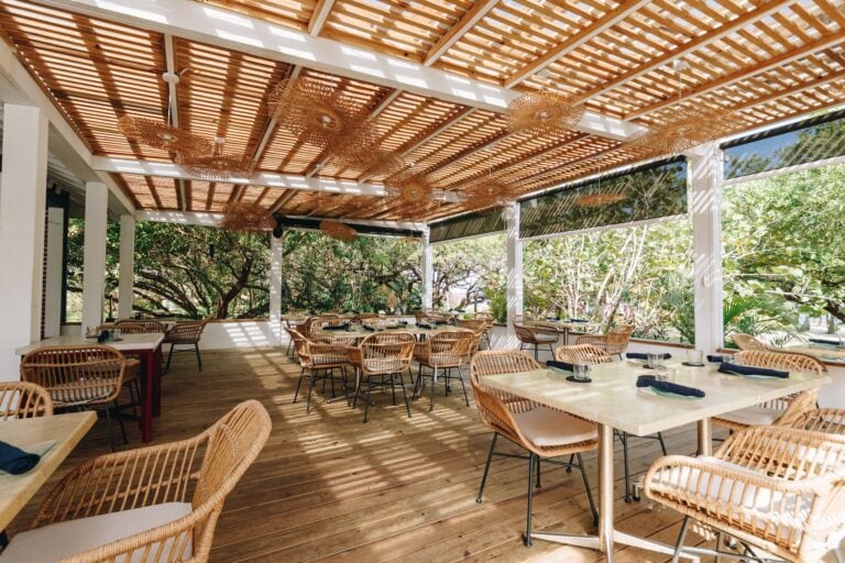 An open-air restaurant in Roatan, Honduras, featuring a wooden slatted ceiling with decorative wicker light fixtures, surrounded by lush tropical greenery and set with tables and rattan chairs.