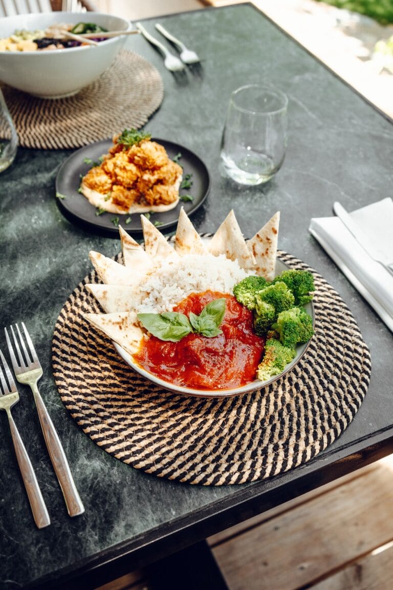A bowl of rice and tomato sauce with broccoli, basil, and tortilla chips is served on a woven placemat at a restaurant in Roatan, Honduras, with a salad and drinking glass on a gray marble table.