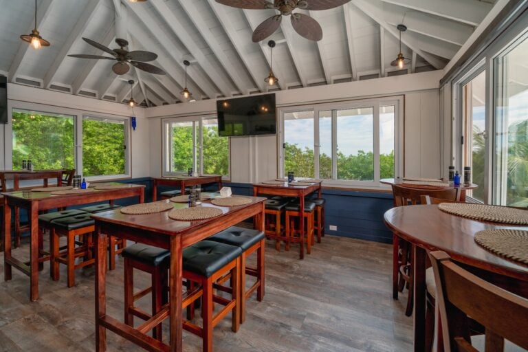An indoor dining area in Roatan, Honduras, features wooden tables with woven placemats, large windows overlooking lush green trees, a white vaulted ceiling with ceiling fans, pendant lights with exposed bulbs, and a mounted flat-screen TV.