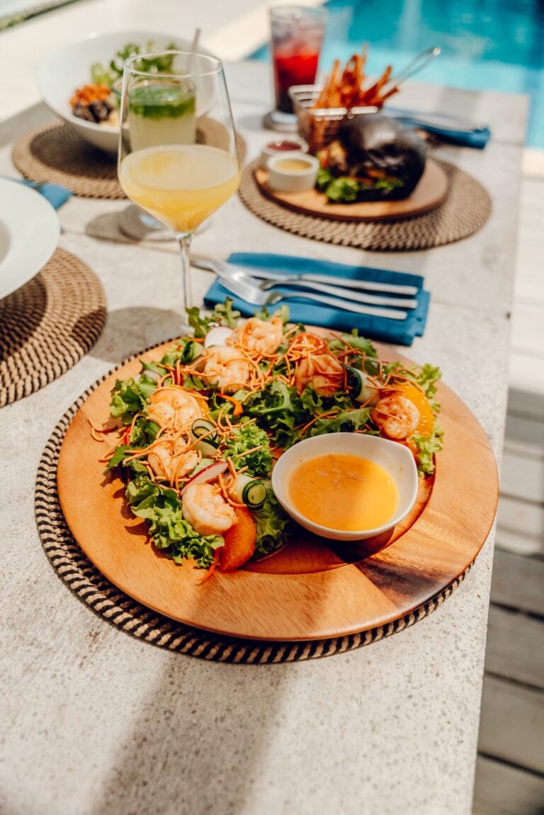 A seafood salad with shrimp, vegetables, and a side of dipping sauce is served on a wooden platter at an outdoor dining area by a pool on Roatan in Honduras.