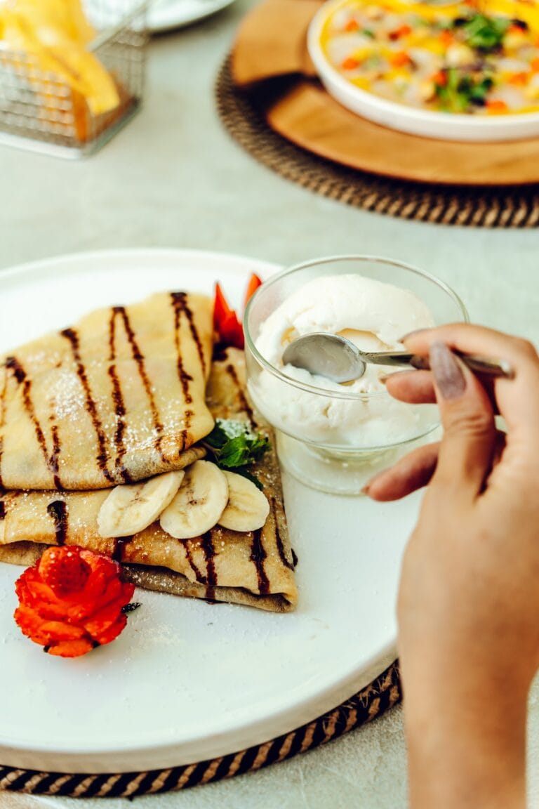 A person is adding whipped cream from a spoon to a glass of banana-flavored dessert served with sliced bananas and strawberries on a white plate, with a tropical breakfast setup including crepes topped with bananas and chocolate syrup in the background on Roatan, Honduras.