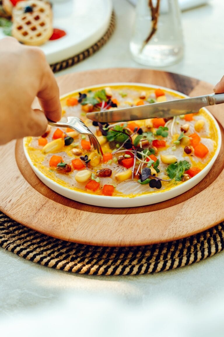 A colorful fish fillet topped with microgreens, vegetables, and herbs, served on a white plate placed on a round wooden tray in a restaurant setting in Roatan, Honduras.