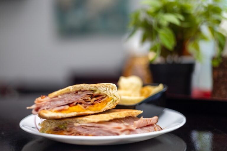 A stacked sandwich with ham, cheese, and lettuce is displayed on a white plate in a dining setting with a blurred background featuring a potted green plant, showcasing the casual culinary atmosphere in Roatan, Honduras.