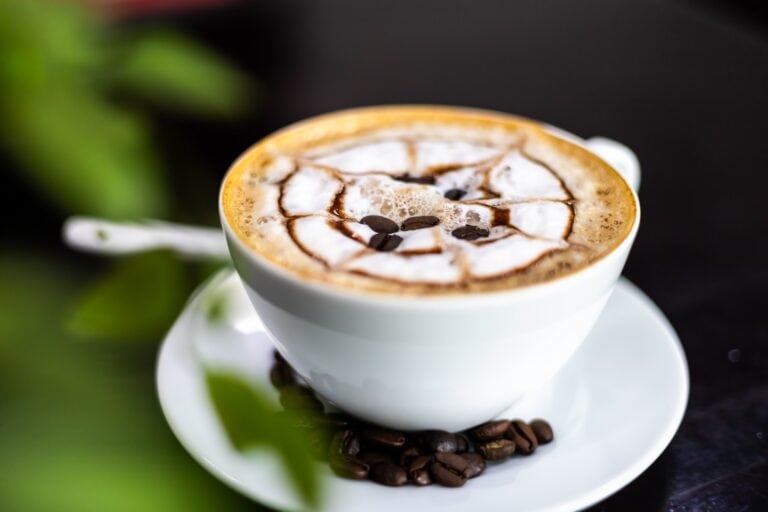 A cup of frothy coffee topped with marshmallows, chocolate drizzle, and coffee beans sits on a saucer, with green leaves in the background, on a dark surface representing a cozy cafe setting in Roatan, Honduras.