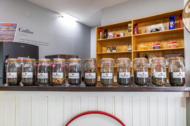 Display of multiple glass jars containing different types of loose leaf teas, arranged on a counter in a coffee shop on Roatan, Honduras, with a wooden shelf of coffee bags and mugs visible in the background.