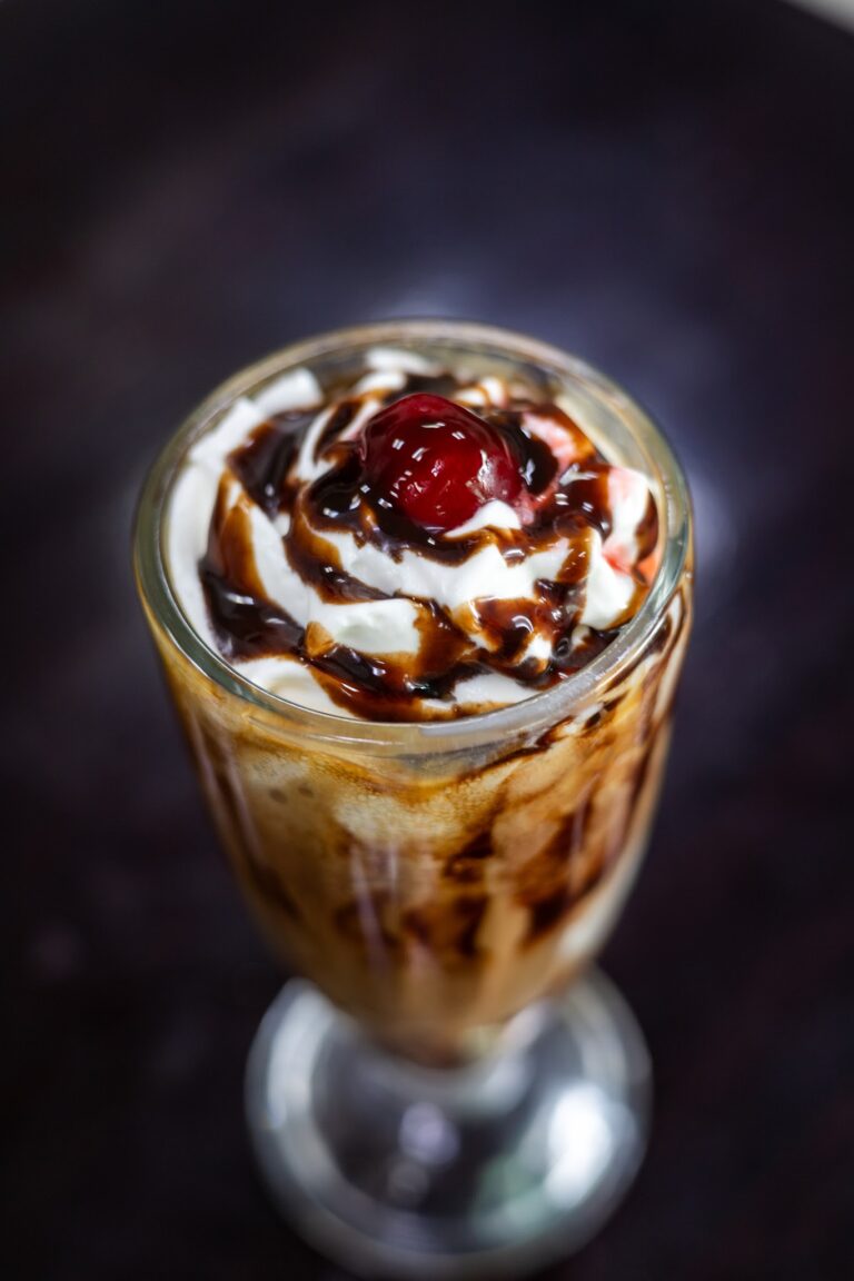 A tall glass of ice cream topped with whipped cream, chocolate syrup, and a maraschino cherry is shown against a black background in this image associated with Roatan, Honduras.