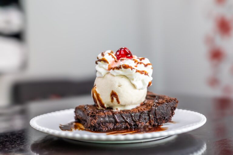 A slice of rich chocolate brownie topped with vanilla ice cream, whipped cream, caramel sauce, and a cherry, served on a white plate against a blurred background in Roatan, Honduras.