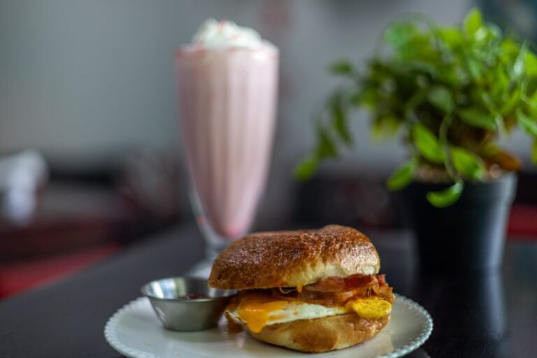 A breakfast sandwich with egg, cheese, and sausage atop toasted bread, served on a white plate with a side of jam or sauce, placed on a dark table with a blurred background of green plants in Roatan, Honduras.
