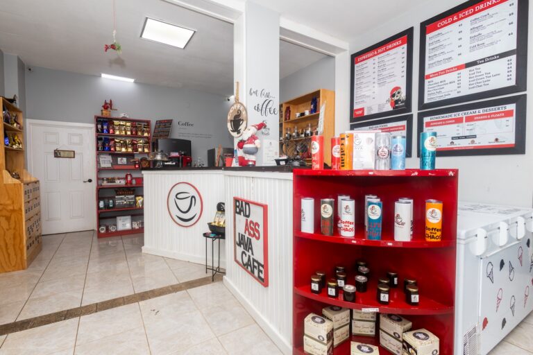 A cozy coffee shop in Roatan, Honduras, features a white counter with red accents, shelves stocked with coffee beans and merchandise, and a menu board displaying various coffee, espresso, tea, and dessert options.