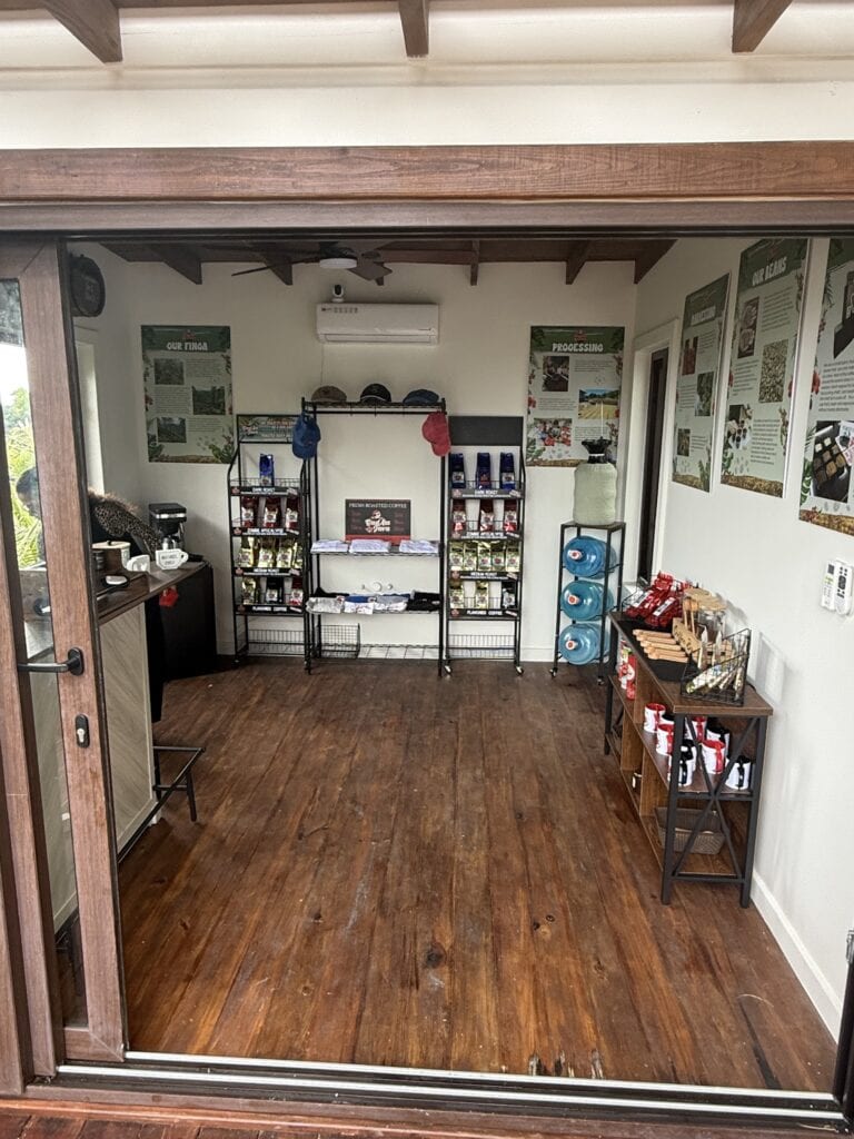 The image shows the interior of a coffee shop or retail space on Roatan, Honduras, featuring display shelves with bags of coffee, informational posters about the coffee processing and farm, and a wooden floor with a ceiling fan and air conditioning unit visible.