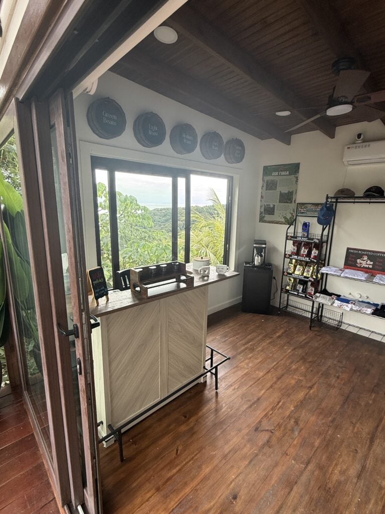 A cozy coffee shop in Roatan, Honduras, features a window with a scenic view of lush greenery and ocean, a display shelf with coffee bags, and decorative roasted coffee signages on the wall.