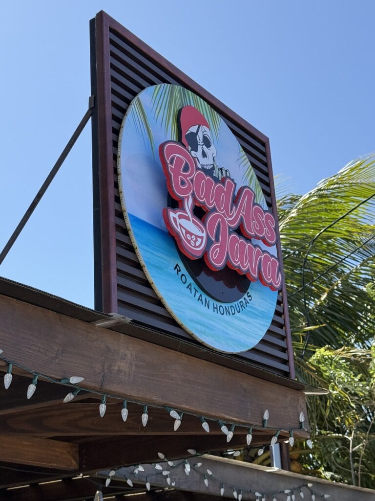A colorful circular sign with a skull wearing a red bandana and sunglasses, surrounded by palm leaves, advertising "Badass Bar" on Roatan, Honduras, hangs above a wooden structure decorated with string lights against a clear blue sky.