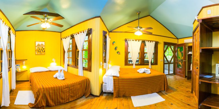 A brightly colored yellow bedroom with two beds, each with white pillows and towel origami, white curtains on wooden windows, and a wooden shelf, located in Roatan, Honduras.