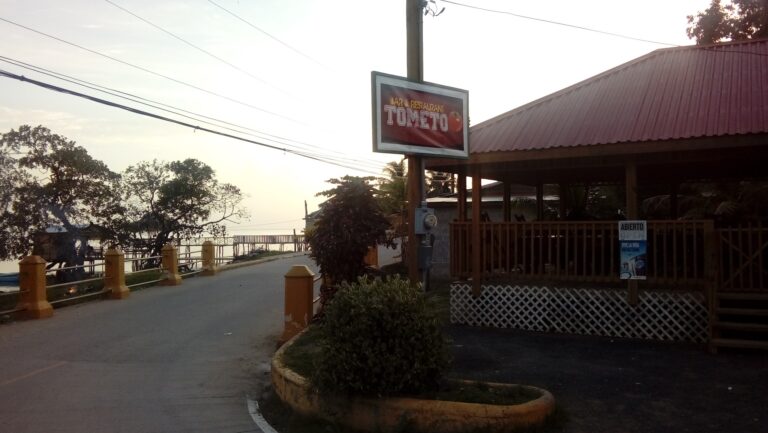 A sign for Tometo bar and restaurant is displayed outside on Roatan, Bay Islands, Honduras, with a wooden building featuring a red metal roof and a waterfront view in the background during sunset.