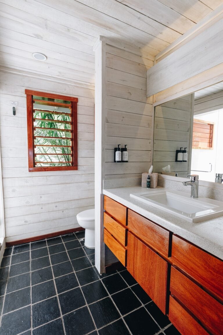 A modern bathroom in Roatan, Honduras features a white wooden wall, a rectangular mirror above a wooden vanity with a white countertop and integrated sink, black tiled flooring, a small window with wooden slats, and black wall-mounted soap dispensers.
