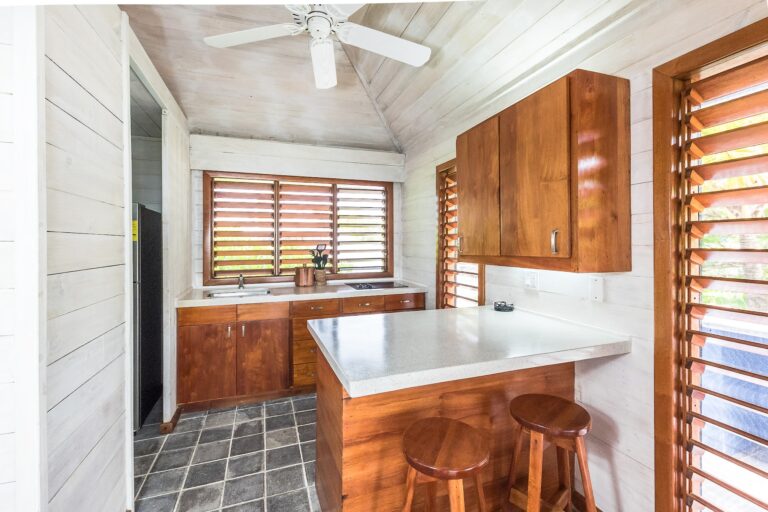 A bright, modern kitchen in a cottage on Roatan, Honduras, features wooden cabinets, a white countertop, louvered wooden windows, and a ceiling fan with a light fixture.