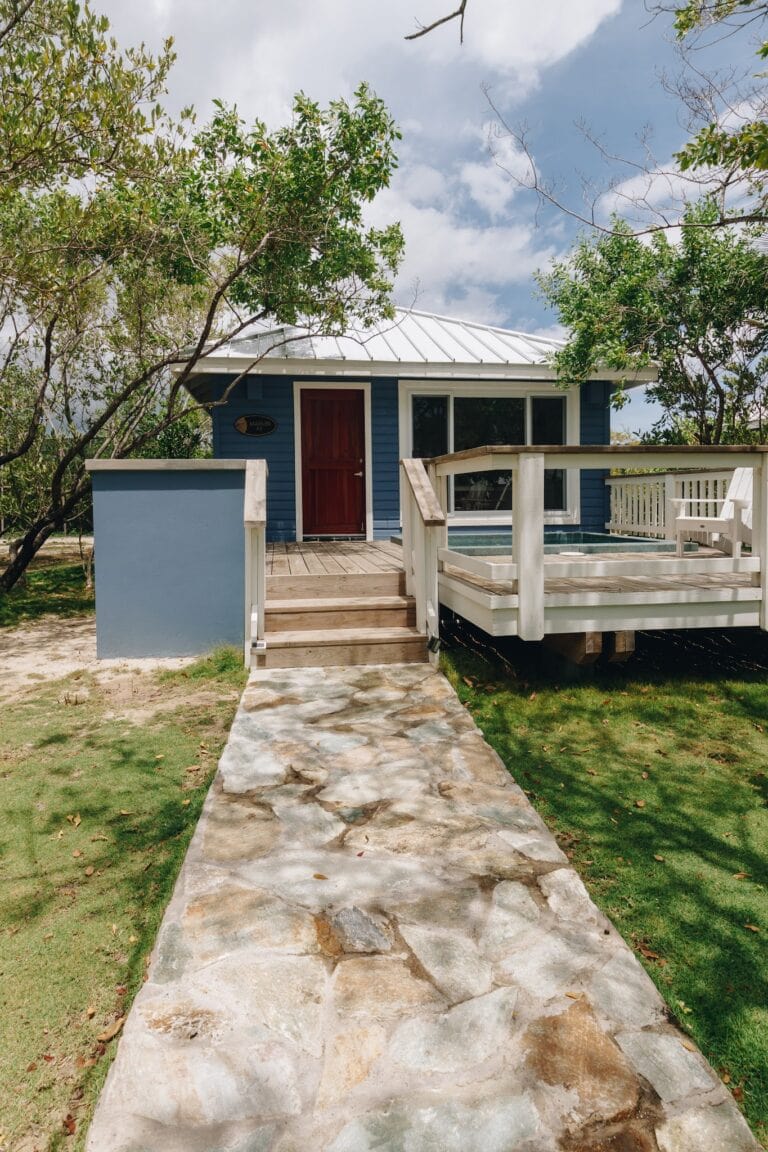 A small blue house with a red door and large windows is situated on a grassy area in Roatan, Honduras, with a stone pathway leading to a wooden deck and surrounded by trees under a partly cloudy sky.