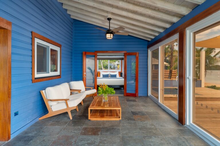 A covered outdoor seating area with a blue wooden wall, white cushioned wooden chairs, a wooden coffee table with a potted plant, large glass sliding doors, and a ceiling fan with a light, set in Roatan, Honduras.