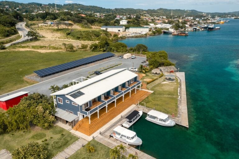 A blue and white waterfront lodging with balconies, located on Roatan in Honduras, features a docked boat, a deck with outdoor seating, and solar panels on the roof, overlooking the harbor and lush hilly landscape.