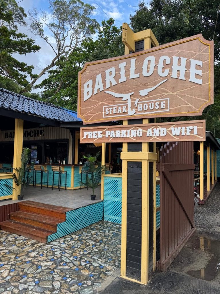 A wooden sign for Bariloche Steak House in Roatan, Honduras, advertises free parking and Wi-Fi, with a view of the restaurant's outdoor seating area and tropical trees in the background.