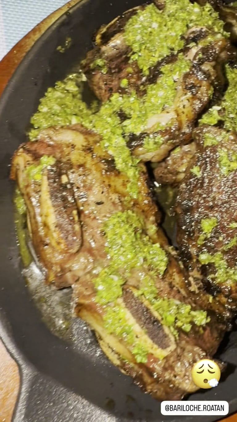 Sliced grilled steak topped with chopped green herbs is shown on a black plate, with a seasoning container in the background, in a dining setting on Roatan, Honduras.