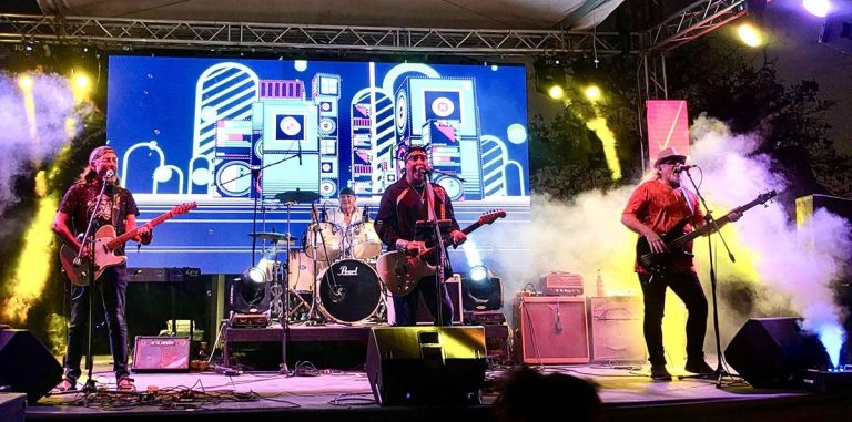 A live band performing on an outdoor stage in Roatan, Honduras, with a colorful animated cityscape projected on a large screen behind them and vibrant yellow and purple stage lighting.