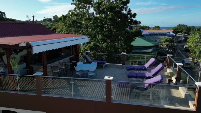A rooftop terrace in Roatan, Honduras, features lounge chairs, a dining area with a table and chairs, and a shaded covered section, offering views of lush greenery and residential buildings in the background.