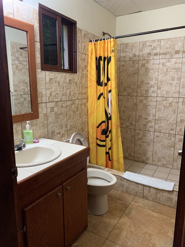 A bathroom with beige tiled walls and floor, featuring a wooden vanity with a sink, a mirror, a window with a wooden frame, a yellow and orange shower curtain, a white towel on the floor, and a toilet in Roatan, Honduras.