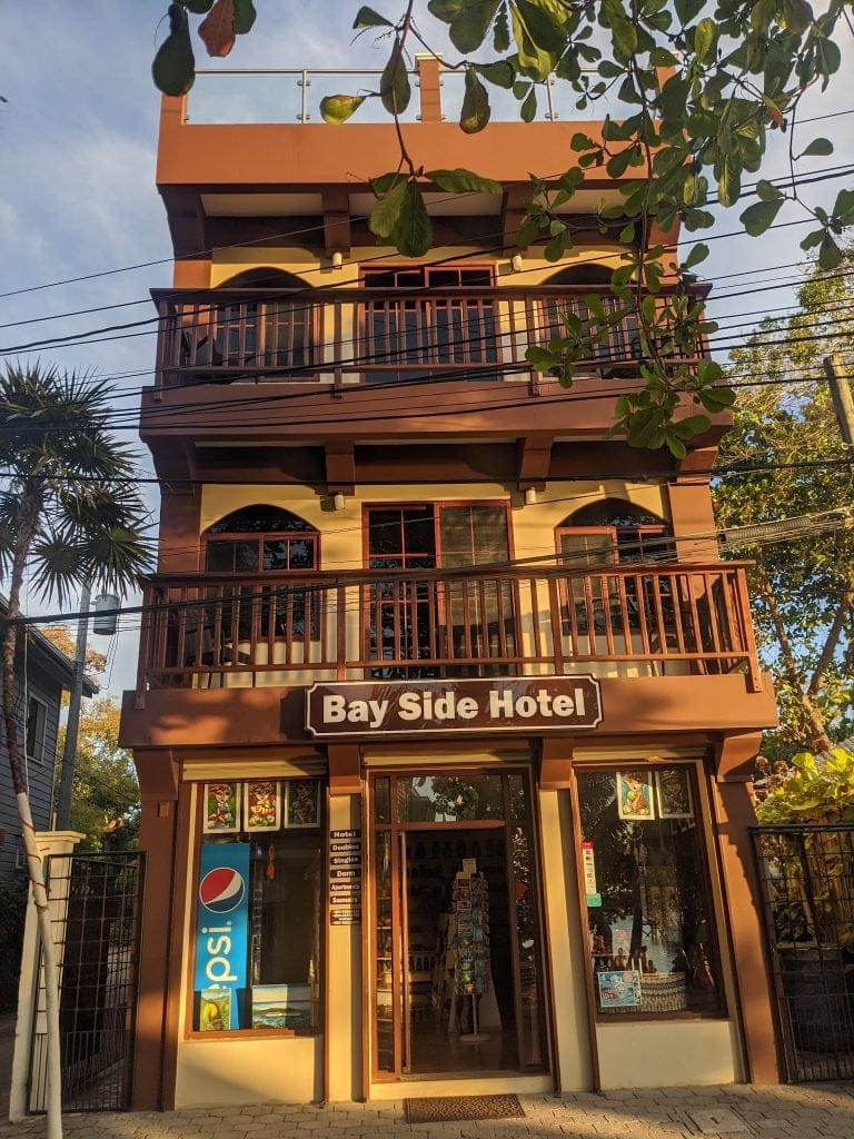 The entrance of the Bay Side Hotel in Roatan, Honduras, features a glass door adorned with colorful artwork, a Pepsi vending machine, and a sign indicating accommodations like doubles, singles, dorms, apartments, and a souvenir shop, with tropical trees partially visible around the building.