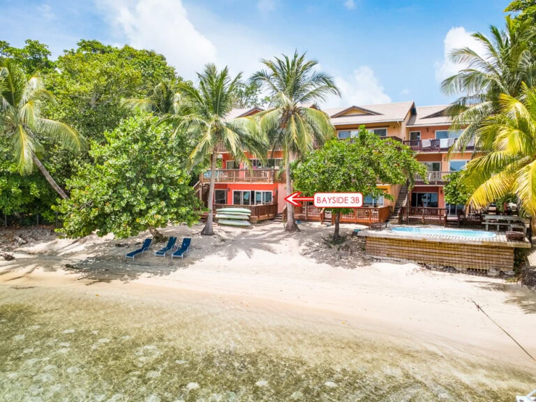 Colorful beachfront building in Roatan, Honduras, with multiple balconies surrounded by palm trees and lush greenery, overlooking a sandy beach with lounge chairs and a small dock with a hot tub.