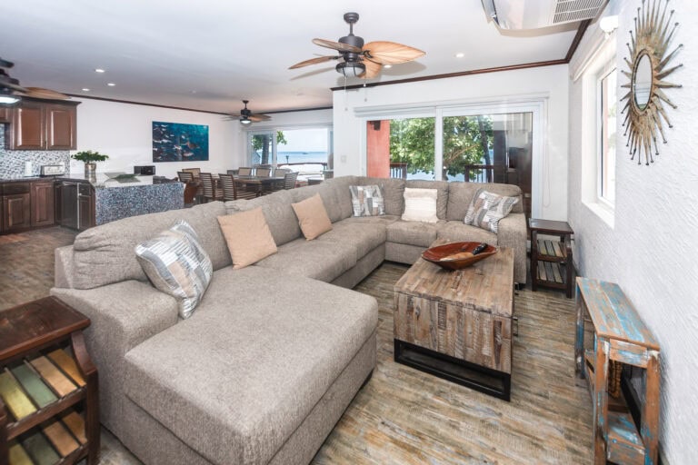 Living room in a beachfront property on Roatan with a large beige sectional sofa, wooden furniture, and sliding glass doors opening to a balcony with ocean views in Honduras.