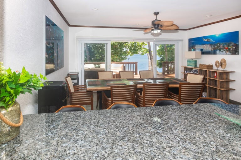 Interior of a dining room in Roatan, Honduras, with a view of a waterfront deck and ocean beyond through glass sliding doors, featuring a large wooden table and tropical decor.